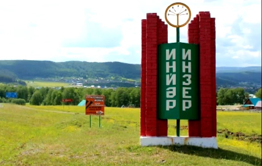 Кирпично-зелёная стела с надписью «Инзер» на холме, внизу село и горы.